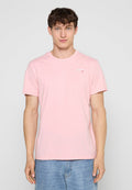 Junger Mann, der ein hellrosa kurzärmeliges T-Shirt und blaue Jeans trägt, vor einem schlichten hellgrauen Hintergrund stehend.