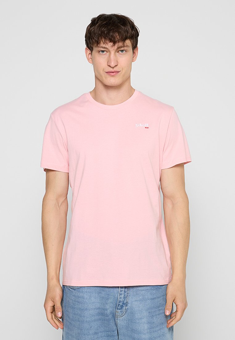 Junger Mann, der ein hellrosa kurzärmeliges T-Shirt und blaue Jeans trägt, vor einem schlichten hellgrauen Hintergrund stehend.