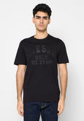 Junger Mann mit kurzen dunklen Haaren trägt ein schwarzes Diesel-T-Shirt mit dem Text "ONLY THE BRAVE", kombiniert mit dunkelblauen Jeans vor weißem Hintergrund.