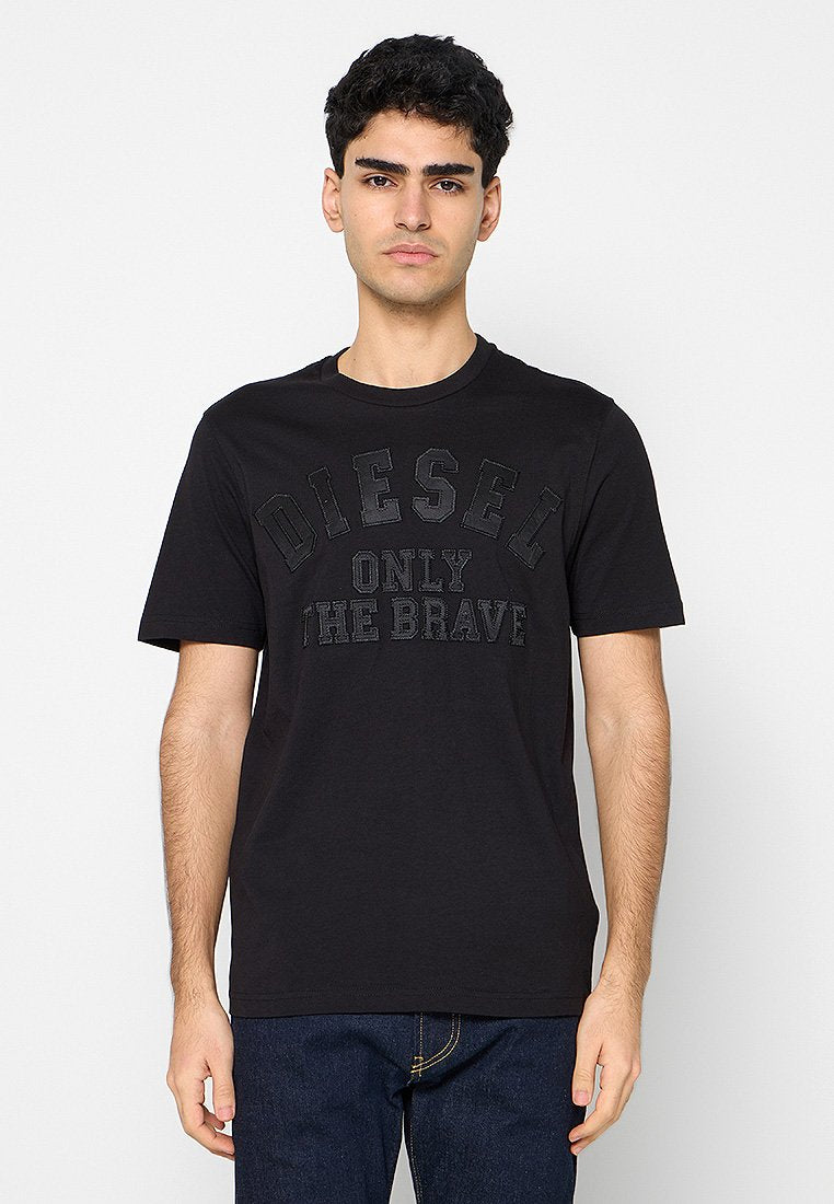 Junger Mann mit kurzen dunklen Haaren trägt ein schwarzes Diesel-T-Shirt mit dem Text "ONLY THE BRAVE", kombiniert mit dunkelblauen Jeans vor weißem Hintergrund.