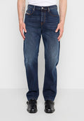 Mann trägt dunkelblaue gerade geschnittene Jeans, weißes Hemd und schwarze Lederschuhe und steht vor einem einfarbigen Hintergrund.