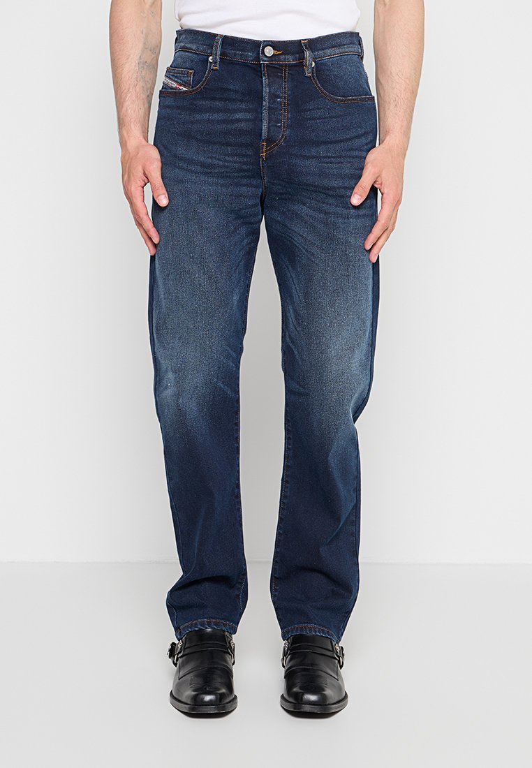 Mann trägt dunkelblaue gerade geschnittene Jeans, weißes Hemd und schwarze Lederschuhe und steht vor einem einfarbigen Hintergrund.