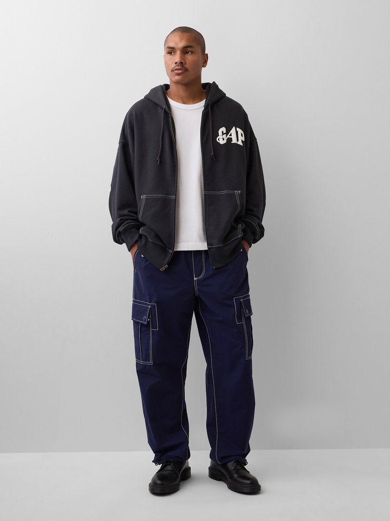 Mann trägt schwarzen GAP Zip-Up-Hoodie, weißes T-Shirt, marineblaue Cargo-Hose mit weißen Nähten und schwarze Schuhe, steht vor einem schlichten grauen Hintergrund.