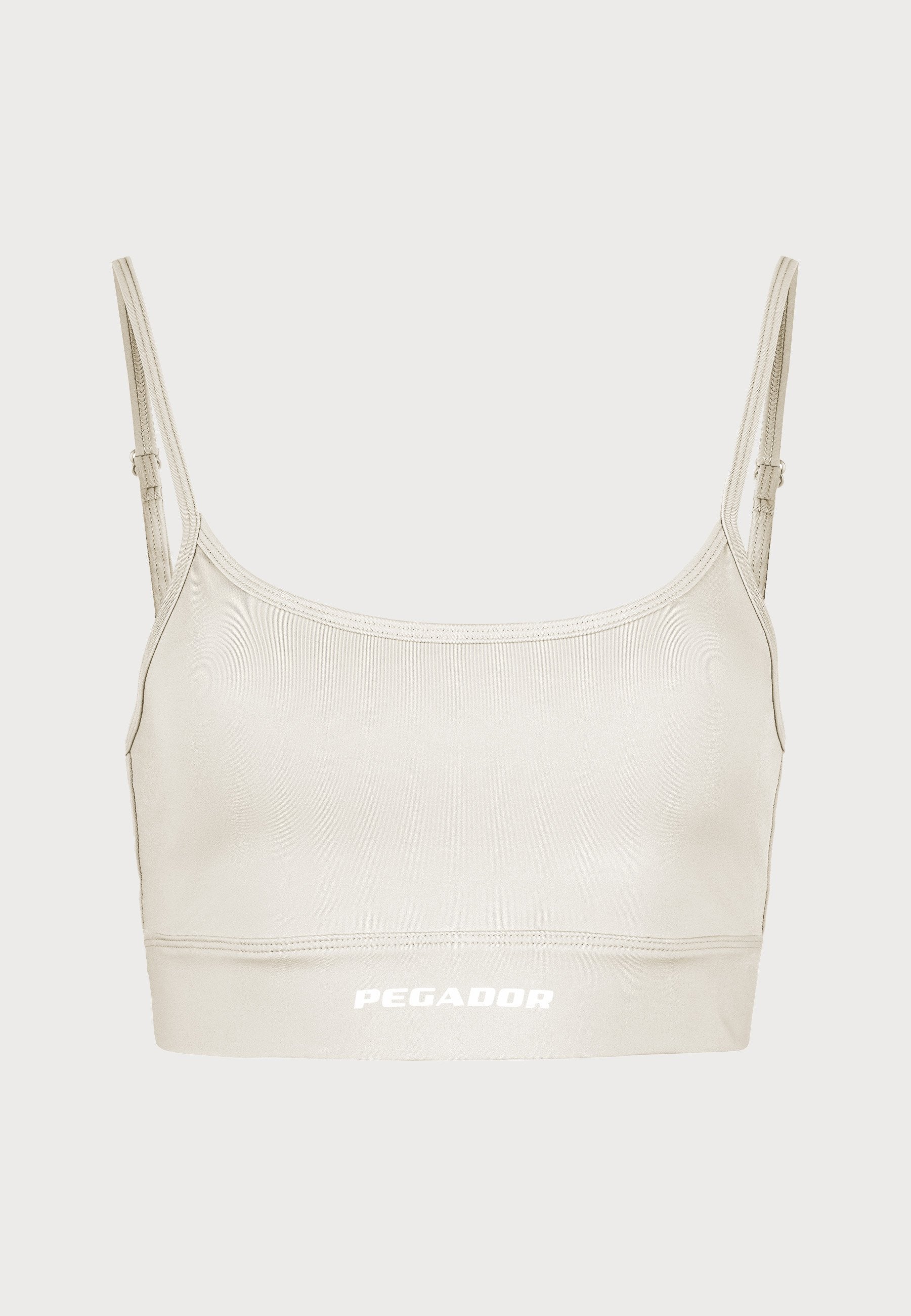 Hellbeige Sport-BH mit dünnen Trägern, schmaler Passform und einem weißen "PEGADOR"-Logo am Saum. Glatte Textur und minimalistischer Stil.