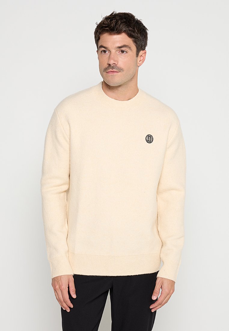 Cremfarbener Pullover aus weichem Stoff, mit Rundhalsausschnitt und geripptem Saum. Kleines schwarzes Logo auf der Brust.