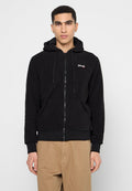Junger Mann in einem schwarzen Reißverschluss-Hoodie mit Fronttaschen und beigen Hosen, der vor einem schlichten hellen Hintergrund steht.