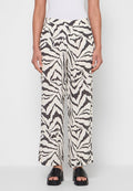 Frau trägt schwarz-weiß abstrakt gemusterte, weit geschnittene Hose, weißes Top und schwarze offene Sandalen mit Absatz, steht vor einfarbigem Hintergrund.