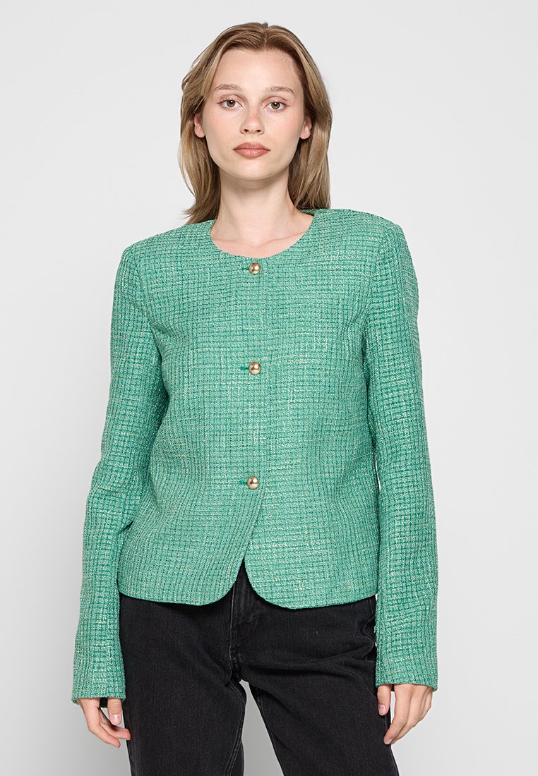 Texturierte grüne Jacke mit rundem Halsausschnitt, drei goldenen Knöpfen, taillierter Passform und langen Ärmeln. Über dunklen Hosen getragen.