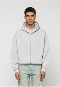 Mann trägt einen hellgrauen Kapuzenpullover mit Reißverschluss und Fronttaschen sowie hellblaue Jeans, steht vor einem schlichten hellen Hintergrund.
