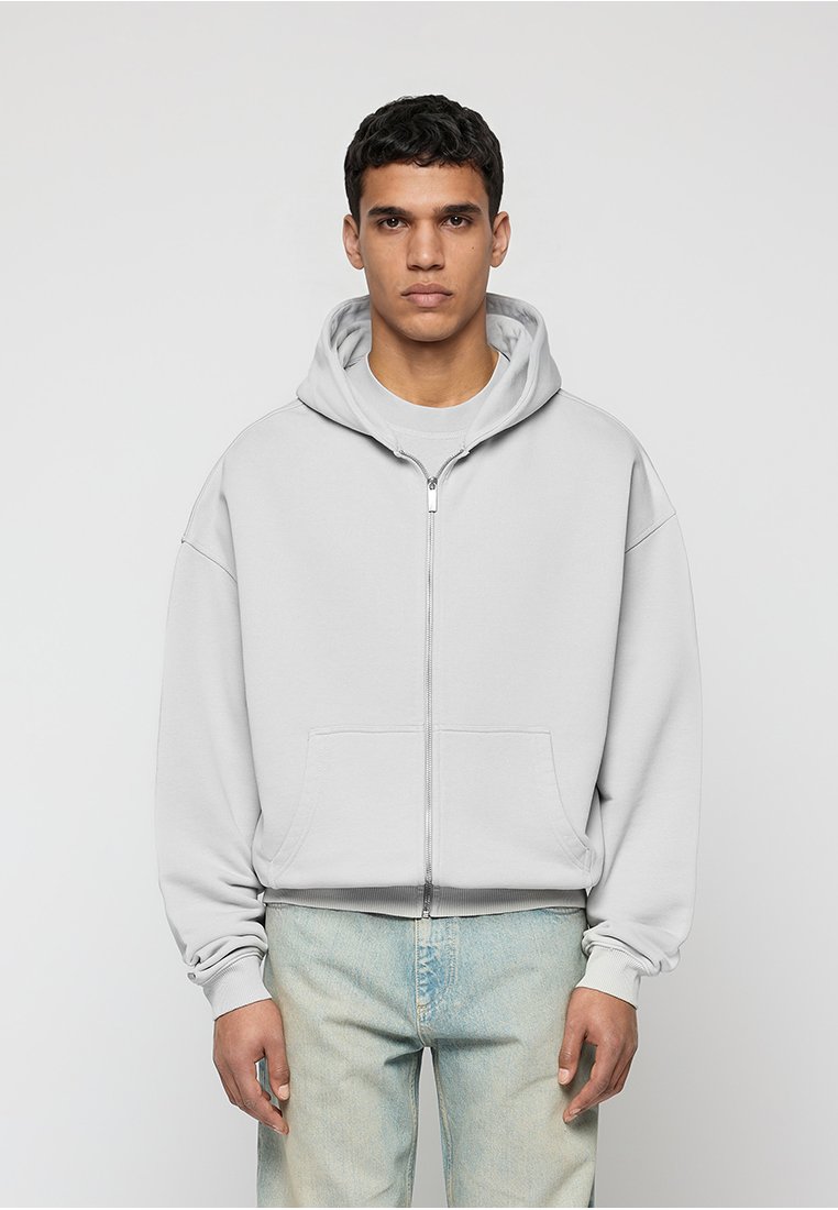 Mann trägt einen hellgrauen Kapuzenpullover mit Reißverschluss und Fronttaschen sowie hellblaue Jeans, steht vor einem schlichten hellen Hintergrund.