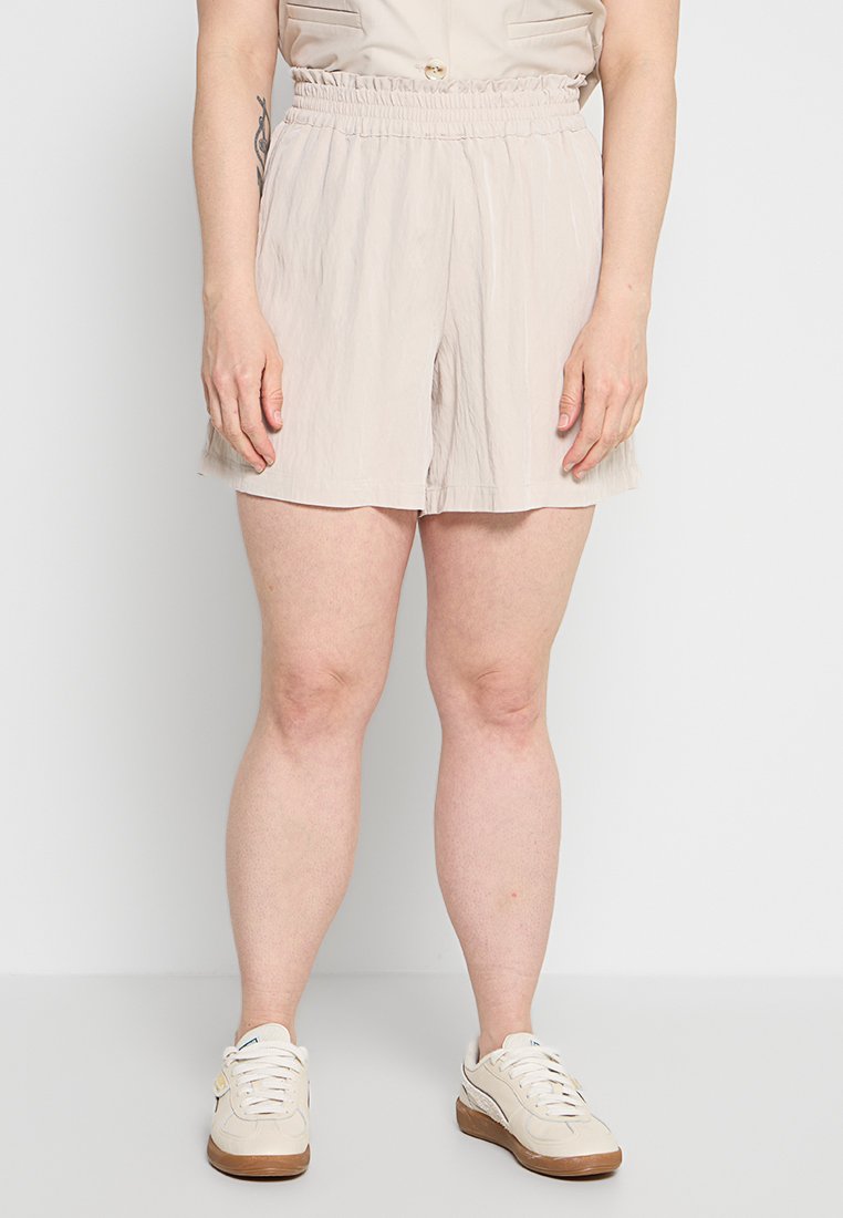 Frau trägt beige Shorts mit elastischem Bund und weiße Turnschuhe, steht vor einem schlichten hellen Hintergrund.