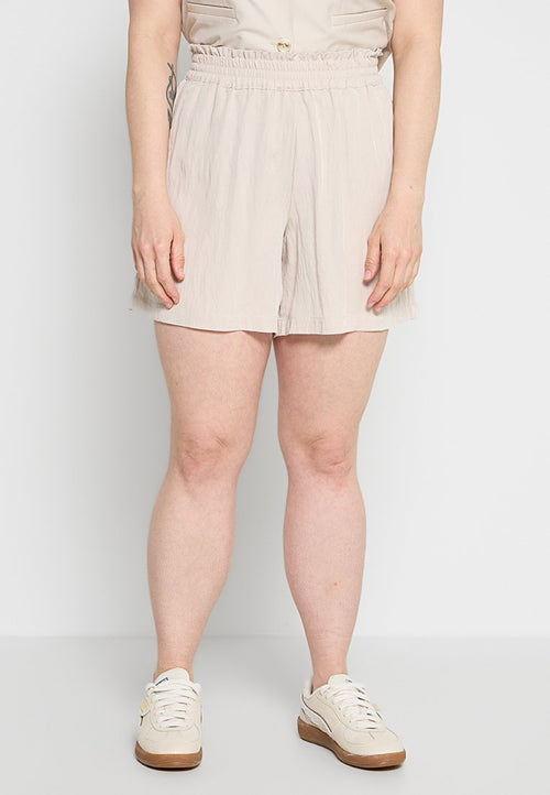 Frau trägt beige Shorts mit elastischem Bund und weiße Turnschuhe, steht vor einem schlichten hellen Hintergrund.