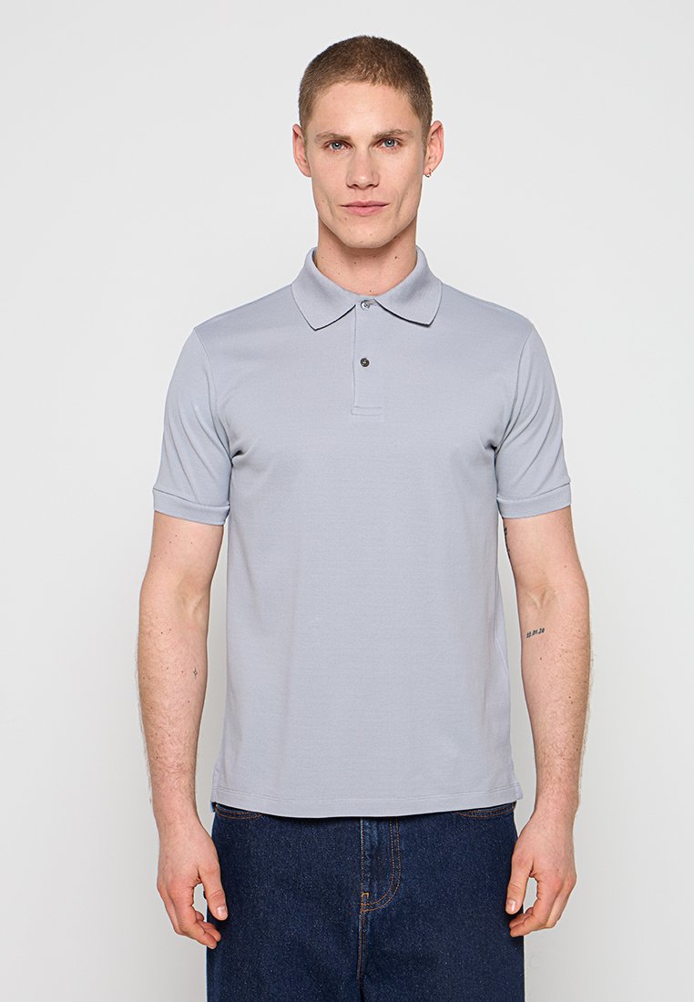 Junger Mann mit kurzen Haaren trägt ein hellgraues Poloshirt mit kurzen Ärmeln und dunkelblaue Jeans, steht vor einem einfachen hellen Hintergrund.