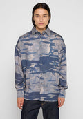 Junger Mann mit langen dunklen Haaren, der ein blau-graues, abstrakt gemustertes Hemd mit Knöpfen und dunkle Jeans trägt, steht vor einem schlichten, hellen Hintergrund.