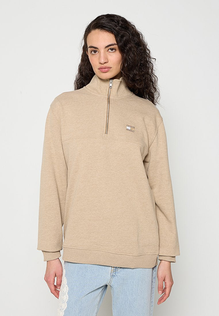Frau mit lockigem dunklem Haar, die einen beigen Sweatshirt mit Viertel-Reißverschluss und hellblaue Jeans mit weißer Spitzenverzierung an der Seite trägt.