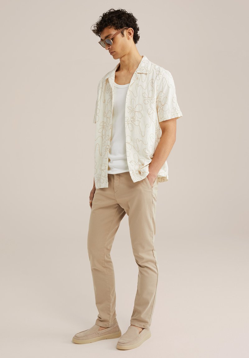 Kurzärmliges cremerfarbenes Hemd mit braunem Blumenmuster, weißes Tanktop, hellbraune Slim-Fit-Hose und beiges Slipper. Stylishe Sonnenbrille rundet den Look ab.