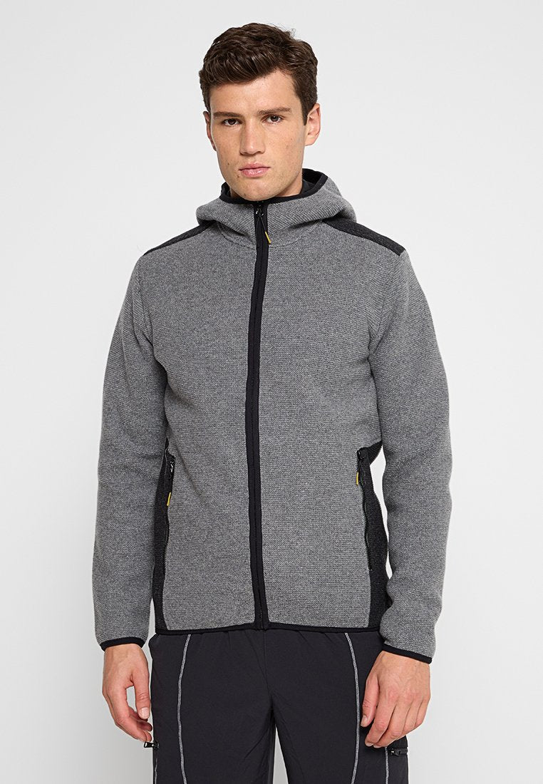 Junger Mann, der einen grauen Reißverschluss-Hoodie mit schwarzen Akzenten und schwarze Hosen trägt, steht vor einem schlichten weißen Hintergrund.