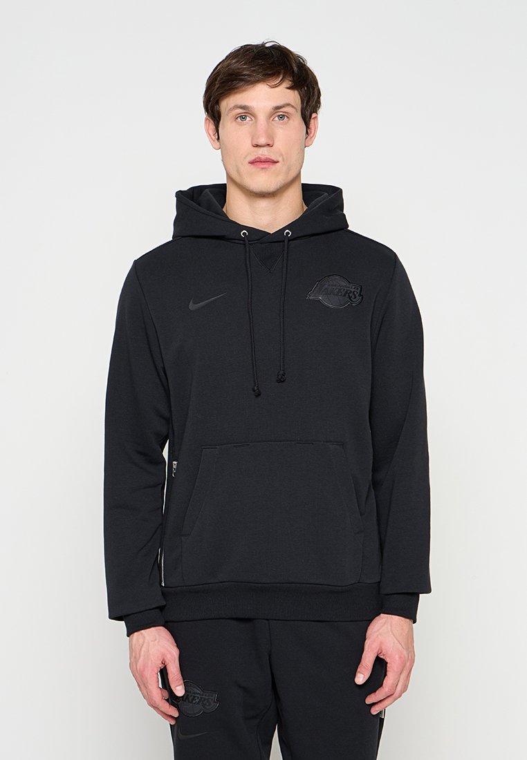 Mann trägt schwarzen Nike-Hoodie und passende Hose mit dezentem dunklem Lakers-Logo, steht vor einem schlichten weißen Hintergrund.