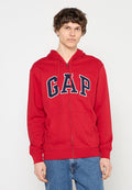 Junger Mann trägt einen roten Kapuzenpullover mit Reißverschluss und dem "GAP"-Logo in Marineblau und Weiß, kombiniert mit blauen Jeans, und steht vor einem schlichten weißen Hintergrund.
