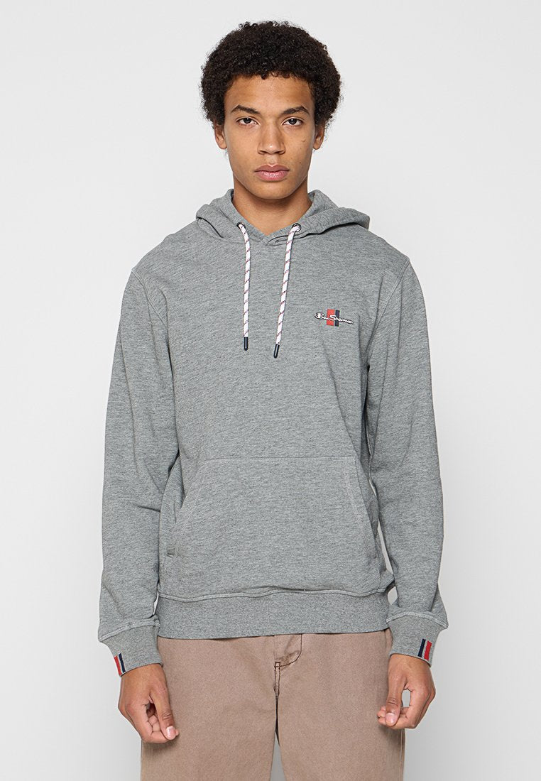 Junger Mann in einem grauen Kapuzenpulli mit weißen Kordeln und beigefarbenen Hosen, der vor einem schlichten hellen Hintergrund steht.