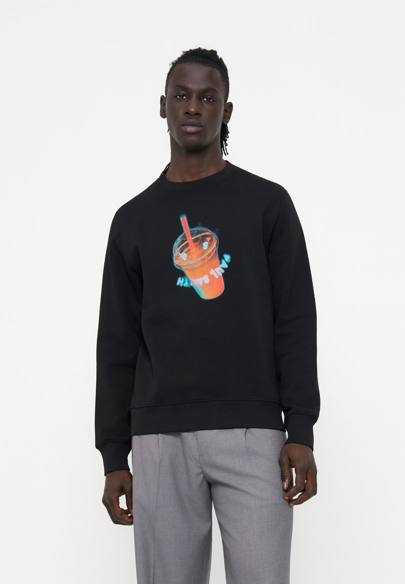 Mann trägt einen schwarzen Sweatshirt mit einer Neon-Style-Grafik eines orangen Getränks mit rotem Strohhalm, kombiniert mit hellgrauen Hosen.