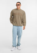 Brauner Oversized-Sweatshirt mit Logo-Print, helle blaue Jeans und weiße Sneaker. Das Modell trägt getönte Sonnenbrillen und steht vor einem schlichten Hintergrund.