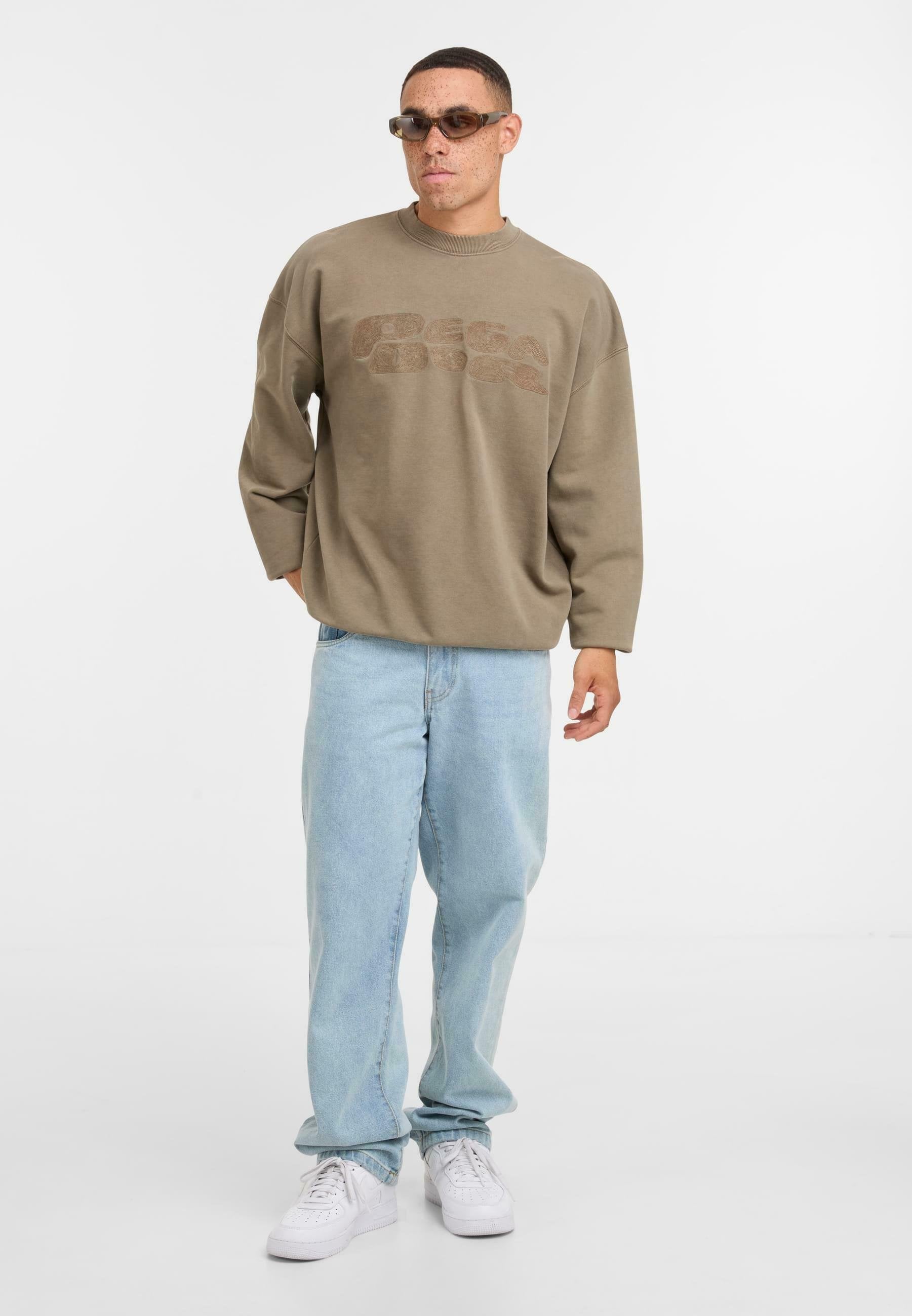 Brauner Oversized-Sweatshirt mit Logo-Print, helle blaue Jeans und weiße Sneaker. Das Modell trägt getönte Sonnenbrillen und steht vor einem schlichten Hintergrund.