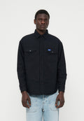 Mann trägt ein schwarzes Hemd mit Knöpfen und Brusttaschen sowie einem kleinen blauen Etikett, kombiniert mit hellblauen Jeans, steht vor einem schlichten Hintergrund.