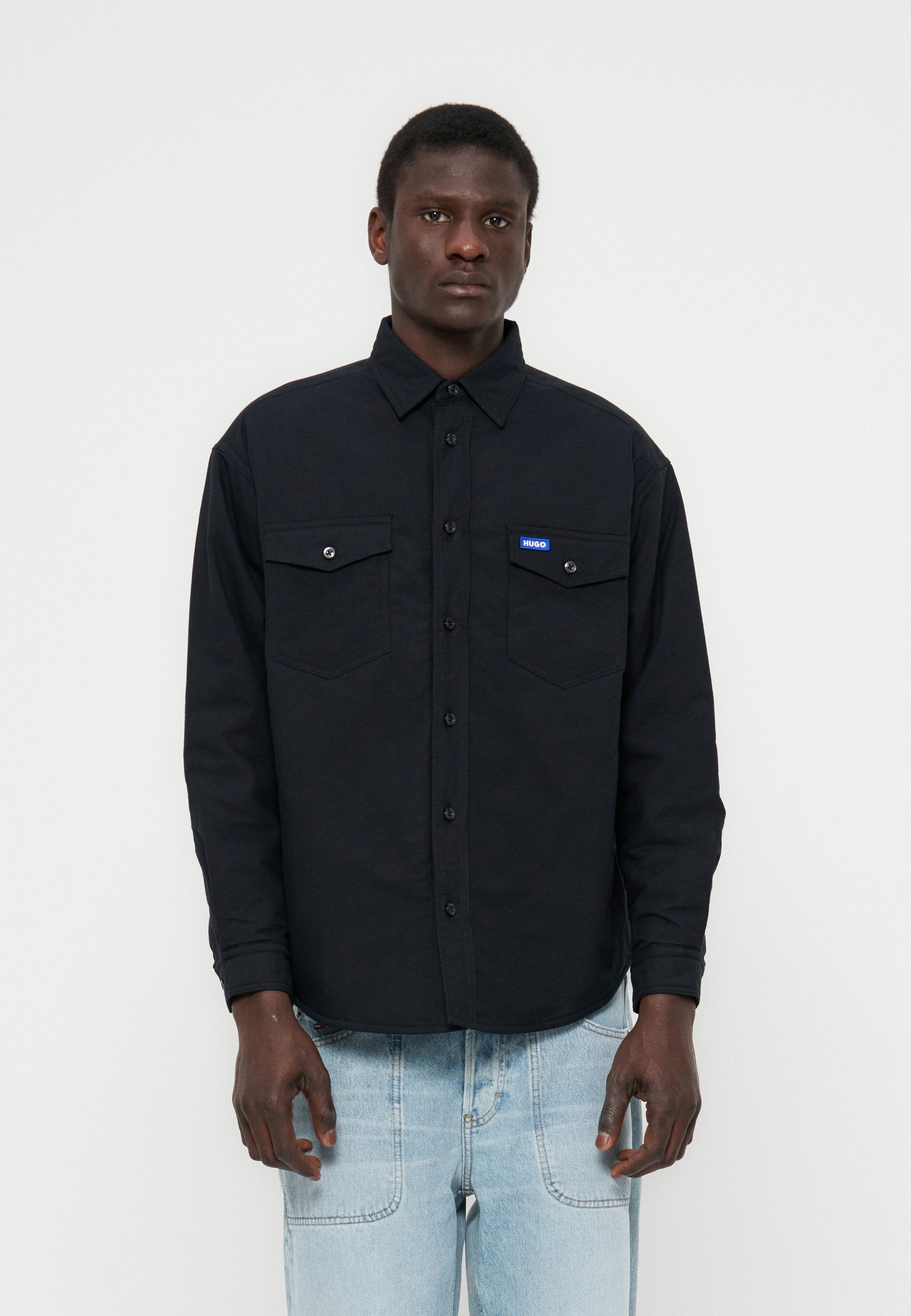 Mann trägt ein schwarzes Hemd mit Knöpfen und Brusttaschen sowie einem kleinen blauen Etikett, kombiniert mit hellblauen Jeans, steht vor einem schlichten Hintergrund.