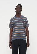 Mann mit dunkler Haut und geflochtenem Haar, der ein graues kurzärmeliges Hemd mit orange-weißen diagonalen Streifen und schwarze Hose trägt.