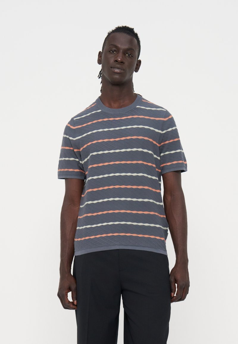 Mann mit dunkler Haut und geflochtenem Haar, der ein graues kurzärmeliges Hemd mit orange-weißen diagonalen Streifen und schwarze Hose trägt.