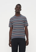 Mann mit dunkler Haut und geflochtenem Haar, der ein graues kurzärmeliges Hemd mit orange-weißen diagonalen Streifen und schwarze Hose trägt.