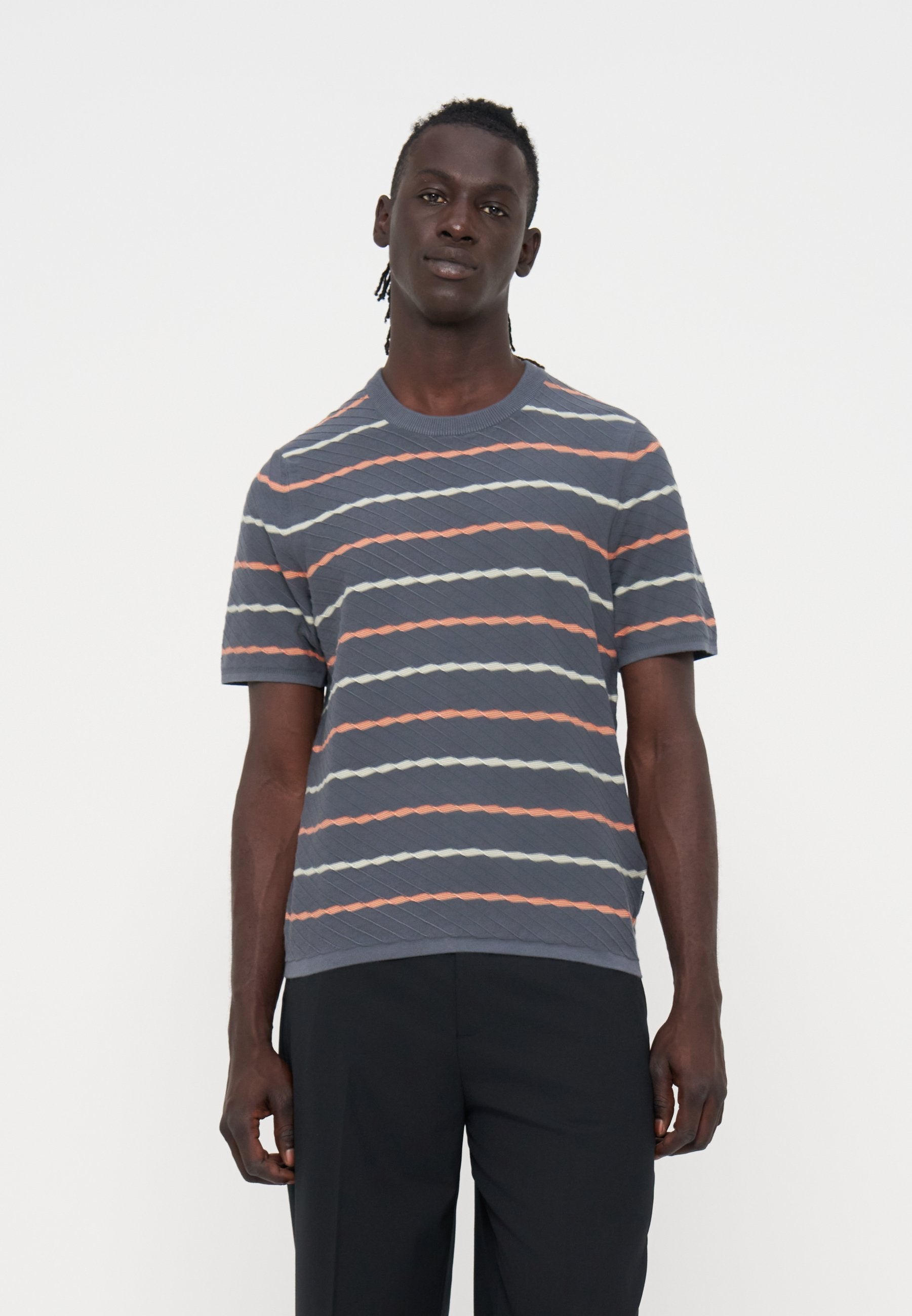 Mann mit dunkler Haut und geflochtenem Haar, der ein graues kurzärmeliges Hemd mit orange-weißen diagonalen Streifen und schwarze Hose trägt.