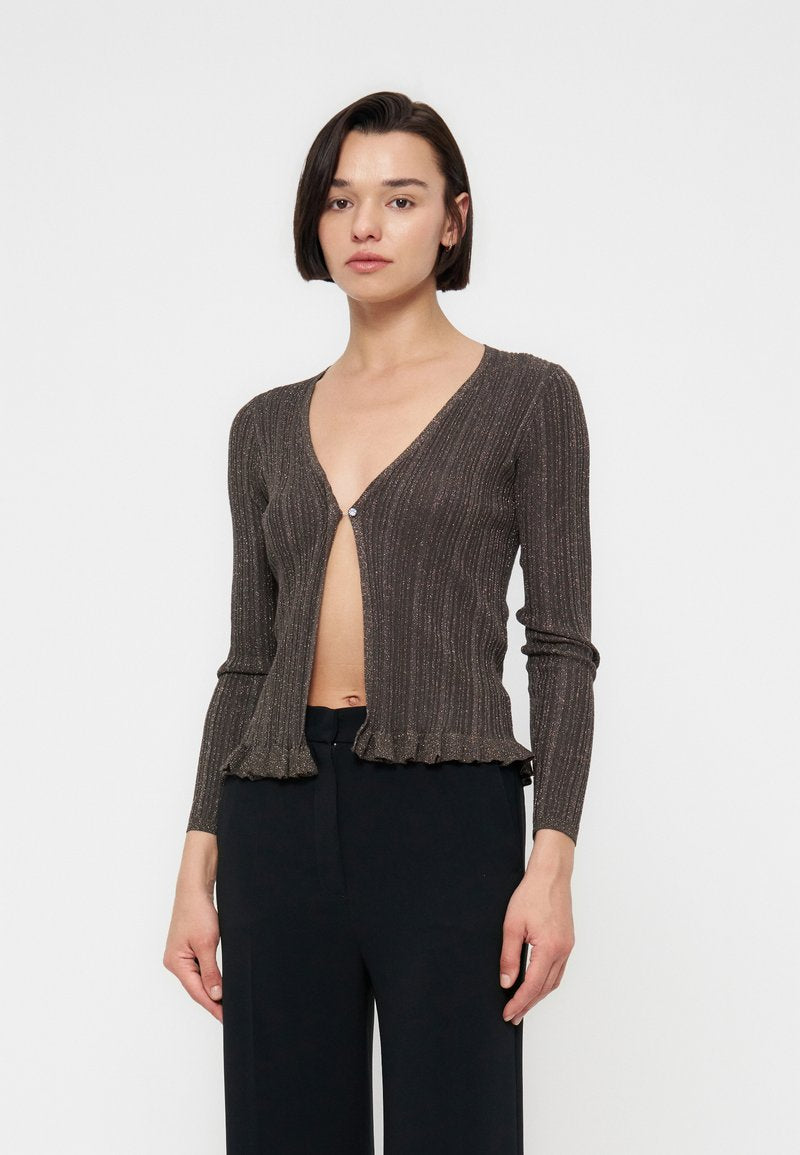 Frau trägt eine braune, gerippte Langarmstrickjacke mit einem Knopf geschlossen, kombiniert mit hoch taillierten schwarzen Hosen vor einfarbigem Hintergrund.