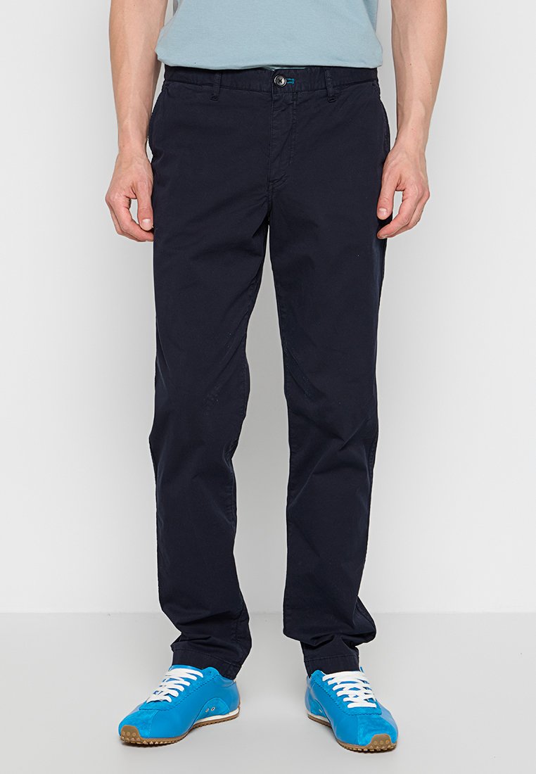 Marineblaue Baumwollhose mit geradem Bein, ausgestattet mit einem vorderen Knopfverschluss und seitlichen Taschen. Kombiniert mit knallblauen Sneakern.