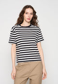 Frau mit langen braunen Haaren, die ein kurzärmeliges, schwarz-weiß gestreiftes Shirt und beige Hosen trägt, steht vor einem schlichten Hintergrund.