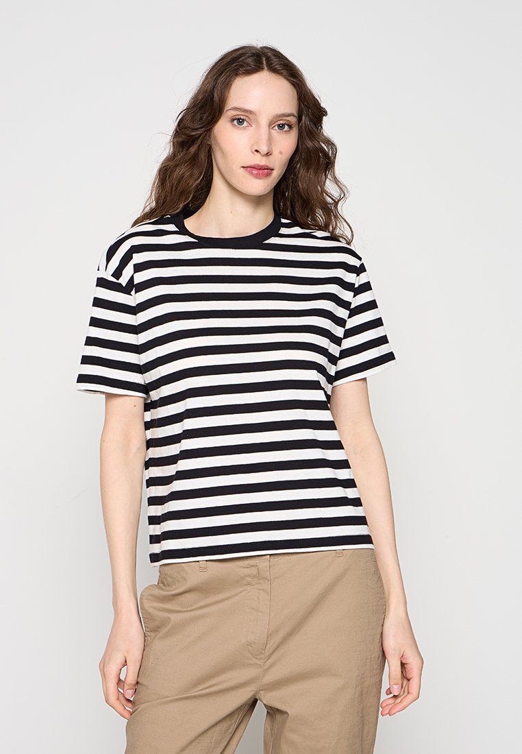 Frau mit langen braunen Haaren, die ein kurzärmeliges, schwarz-weiß gestreiftes Shirt und beige Hosen trägt, steht vor einem schlichten Hintergrund.