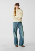 Hellecremefarbener, gestrickter Pullover mit halbem Reißverschluss; lockere blaue Jeans; braune Mütze mit weißem Text; braune spitze Stiefel; glatte Stofftextures.