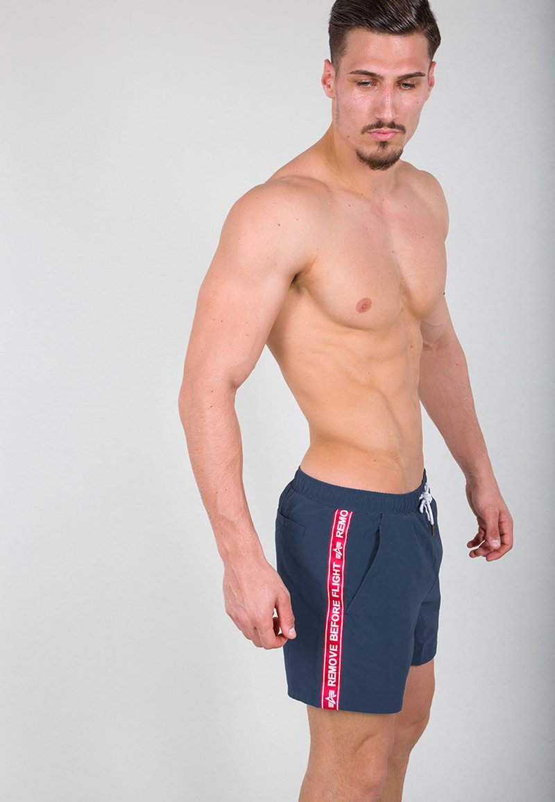 Marineblaue Schwimmshorts mit elastischem Bund und Kordelzug, ausgestattet mit einem roten Seitenstreifen, auf dem "REMOVE BEFORE FLIGHT" steht.