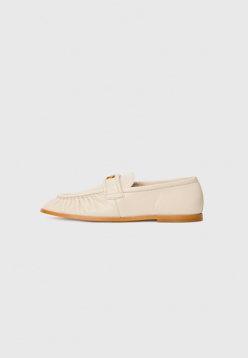 Helle beige Lederloafers mit runder Zehenform, gefälteltem Detail und einem goldenen Akzent an einem Riemen. Flache Sohle mit glatter Textur.