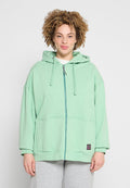 Person mit lockigem Haar, die eine mintgrüne Kapuzenjacke mit Reißverschluss und hellgraue Hose trägt und vor einem schlichten hellen Hintergrund steht.