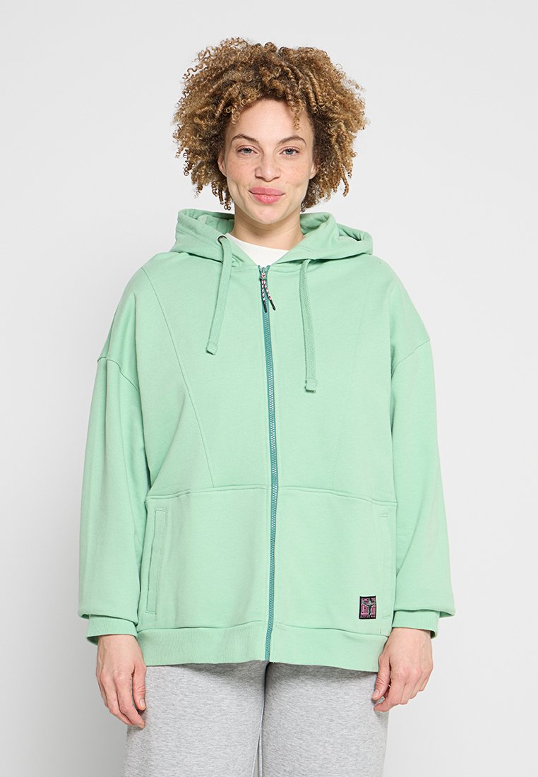 Person mit lockigem Haar, die eine mintgrüne Kapuzenjacke mit Reißverschluss und hellgraue Hose trägt und vor einem schlichten hellen Hintergrund steht.