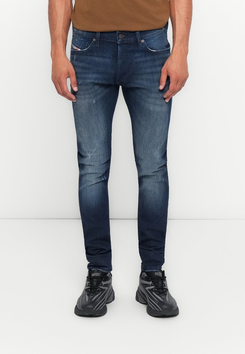 Mann in schmal geschnittenen blauen Jeans und schwarzen Sneakers, der vor einem schlichten weißen Hintergrund steht.
