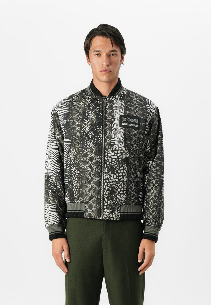 Mann in einer schwarz-weiß gemusterten Bomberjacke mit Tiermotiven und grüner Hose, der vor einem schlichten weißen Hintergrund steht.