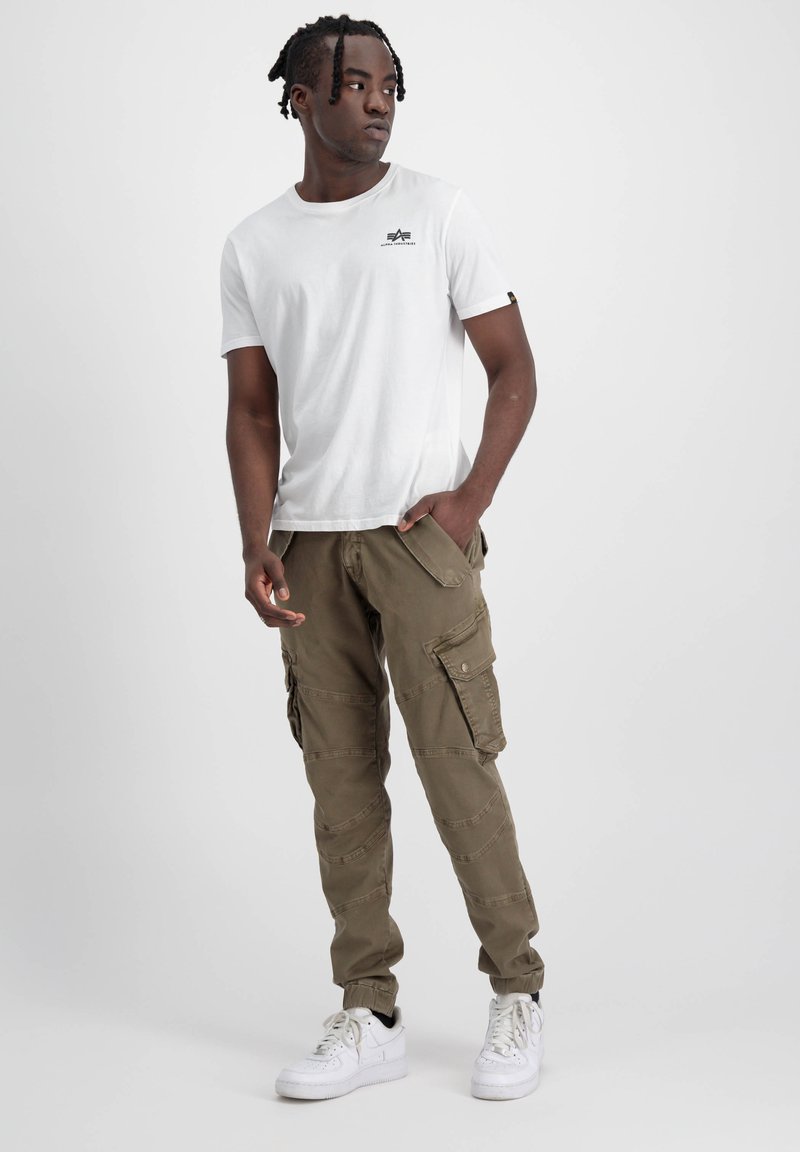 Weißes T-Shirt mit kurzen Ärmeln und kleinem schwarzem Logo, kombiniert mit olivgrünen Cargo-Hosen mit mehreren Taschen und schmalem Schnitt. Weiße Sneakers.