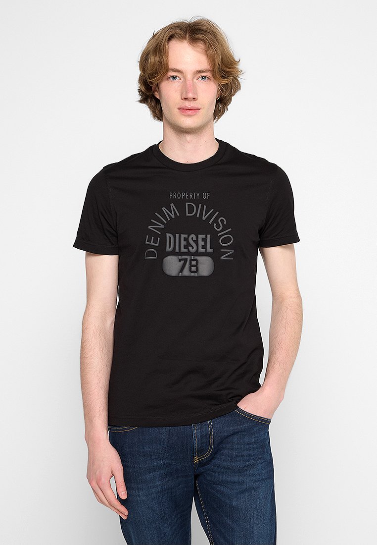 Junger Mann mit hellbraunen Haaren, der ein schwarzes Diesel-T-Shirt mit dem Aufdruck "Denim Division 78" und blaue Jeans trägt und mit einer Hand in der Tasche steht.