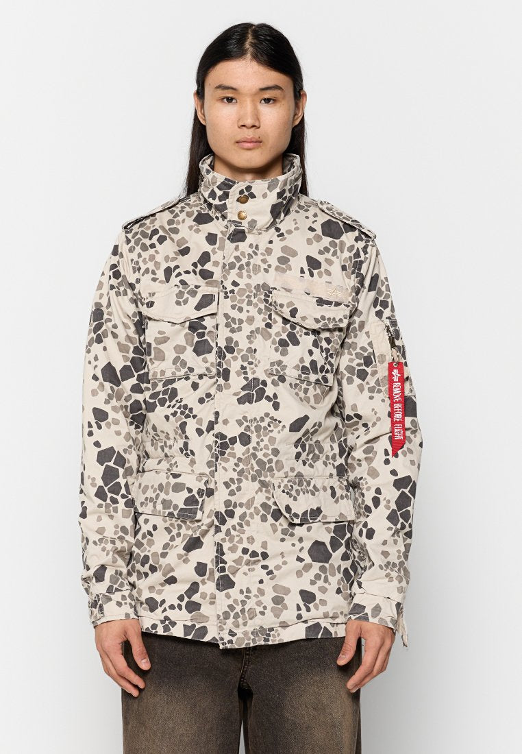 Tarnmusterjacke in Beige mit schwarzen und grauen Akzenten. Mit einem hohen Kragen, vier Fronttaschen und einem roten Anhänger am Sleeve.