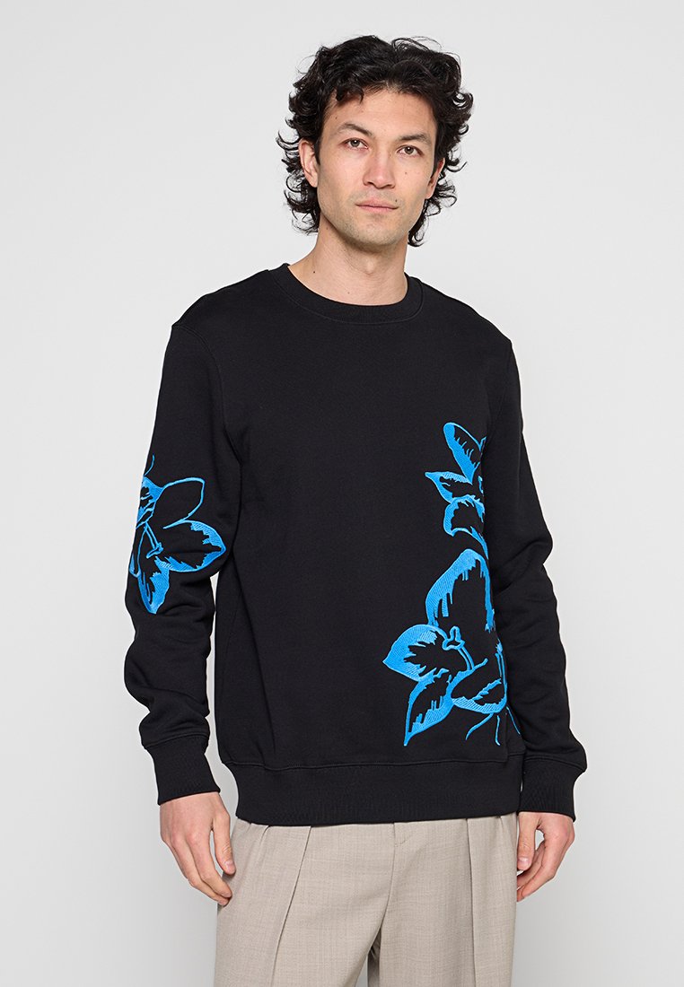 Mann trägt schwarzen Pullover mit großen blauen Blumenmustern auf Brust und linkem Ärmel, kombiniert mit beigen Hosen, steht vor weißem Hintergrund.