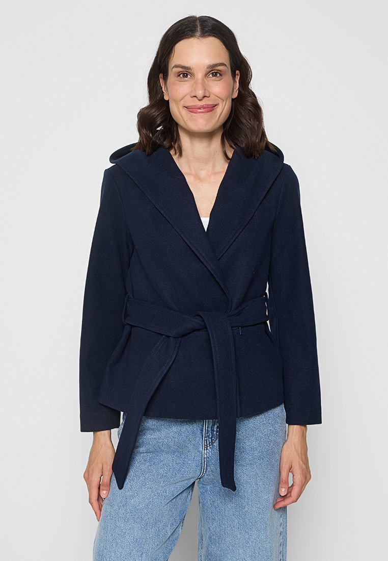Marineblaue Wickeljacke mit großem Kragen, tailliertem Gürtel und langen Ärmeln, aus weichem Material gefertigt. Kombiniert mit hellen Jeans.