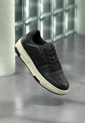 Schwarzer Ledersneaker mit weißer Sohle und schwarzen Schnürsenkeln, der über einem grauen Boden und einer Wand aus Glasbausteinen schwebt.